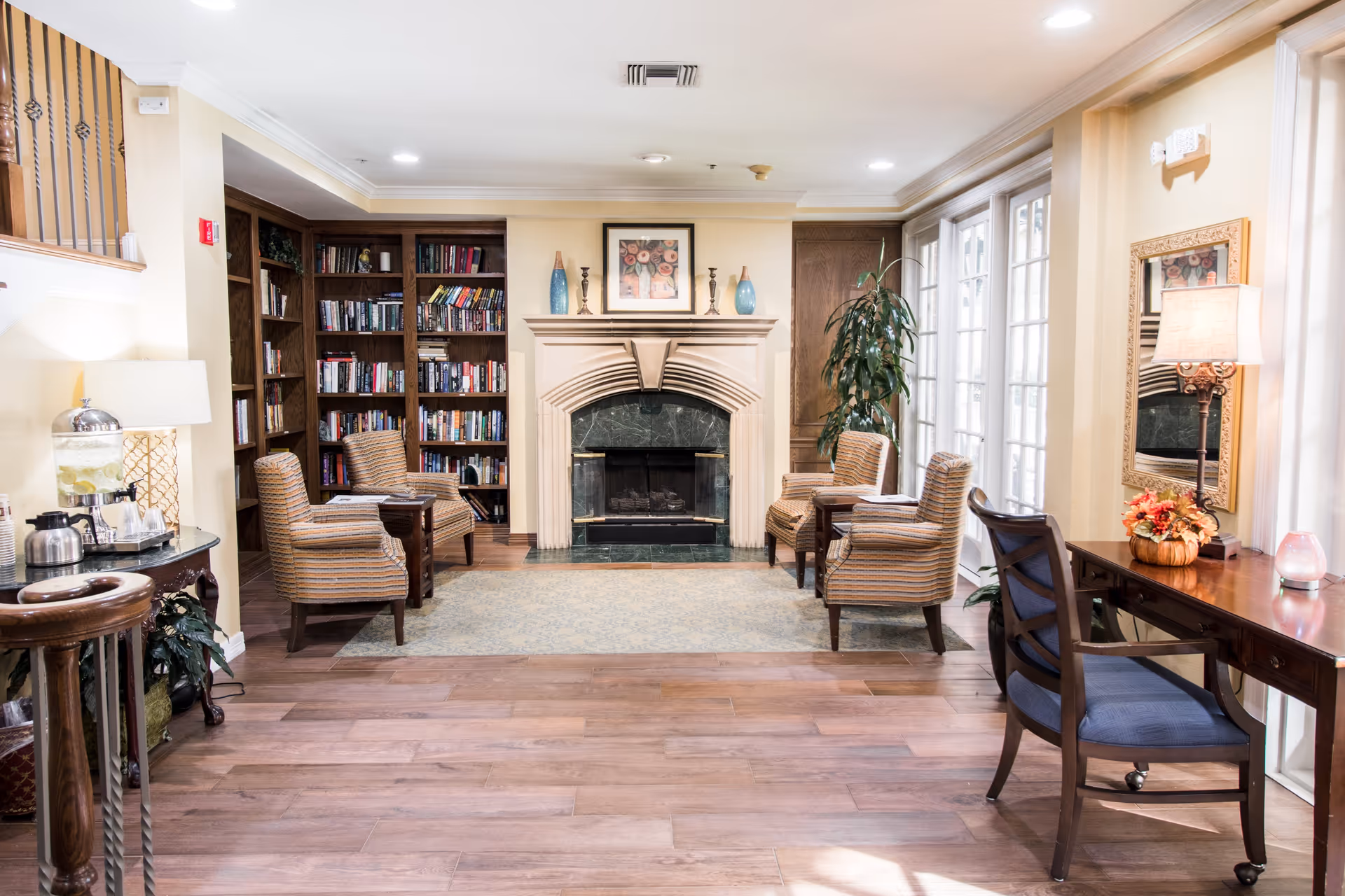 A cozy living room area with a fireplace centered on the back wall, flanked by built-in bookshelves filled with books. Four striped upholstered chairs are arranged around small wooden tables on a light blue patterned rug. To the right, there is a wooden desk with a blue cushioned chair, a table lamp, and a small floral arrangement. Large windows with white frames allow natural light to fill the room. The floor is wood with a warm tone.