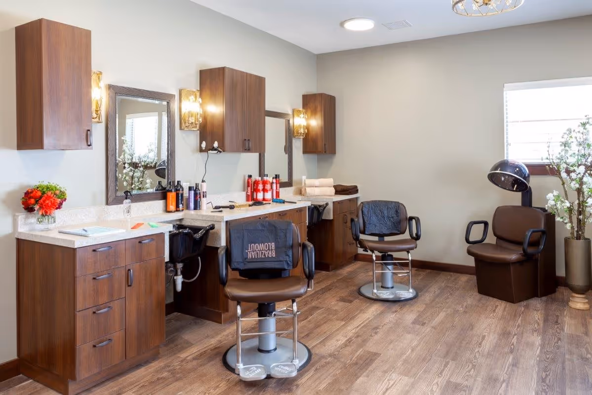 Interior of a salon area with two brown salon chairs in front of mirrors and wooden cabinets. Various hair care products and towels are placed on the countertops. A hair dryer chair is positioned near a window with a potted plant beside it. The room has wooden flooring and soft lighting.