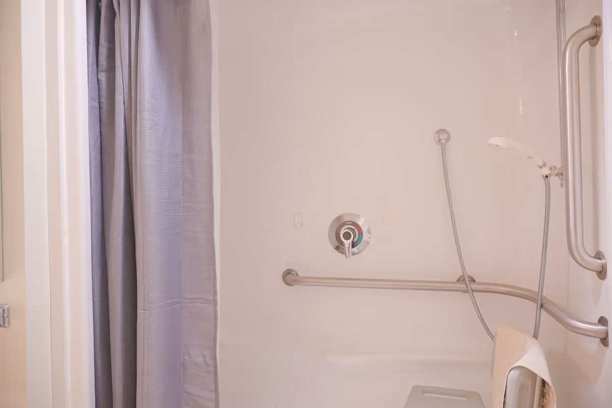 Interior view of a bathroom shower area with a gray shower curtain, a handheld showerhead, grab bars, and a foldable shower seat with a towel on it.