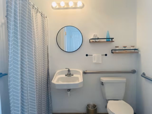 Small accessible bathroom with sink, toilet, grab bars, round mirror, floating shelves, and a shower with a patterned curtain.