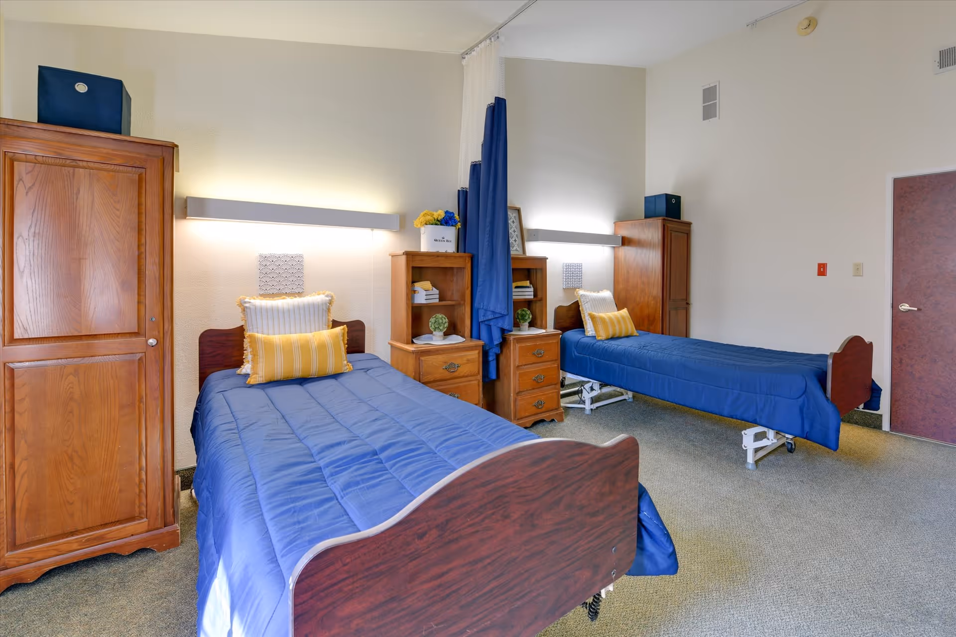 A senior living facility bedroom with two single beds, each covered with blue bedspreads and yellow accent pillows. Between the beds is a wooden nightstand with drawers and a small shelf holding decorative items. Each bed has a wooden headboard and footboard. There are two wooden wardrobes, one near each bed, and a blue privacy curtain hanging between the beds. The room has beige walls and carpeted flooring.