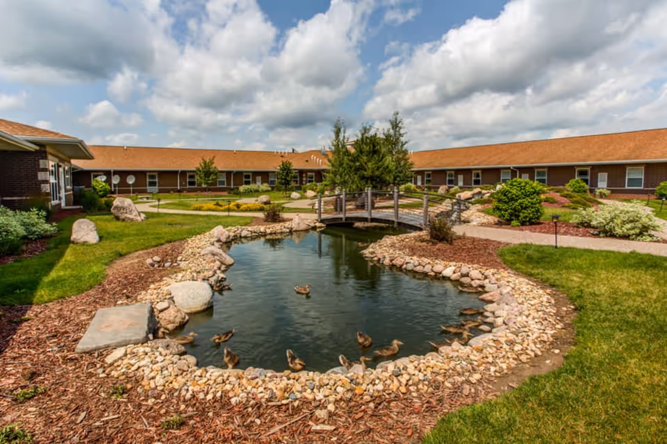 Outdoor garden area at Addington Place of Clinton featuring a small pond with ducks, a wooden footbridge, landscaped greenery, bushes, and a building with a brown roof in the background under a partly cloudy sky.