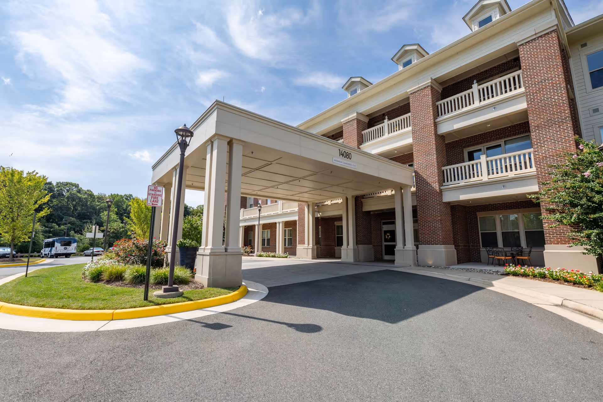 Front entrance of a multi-story brick building with white columns and balconies, a covered driveway area, landscaped greenery, and a clear blue sky.