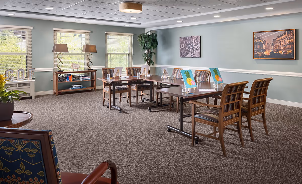 A well-lit room with large windows, light green walls, and carpeted floor. There are two tables arranged in a row with wooden chairs around them. On the tables are small easels holding paintings of pineapples and glasses of water. A wooden console table with two lamps and books is against the wall under the windows. Two framed pictures hang on the walls, and a tall potted plant is in the corner.