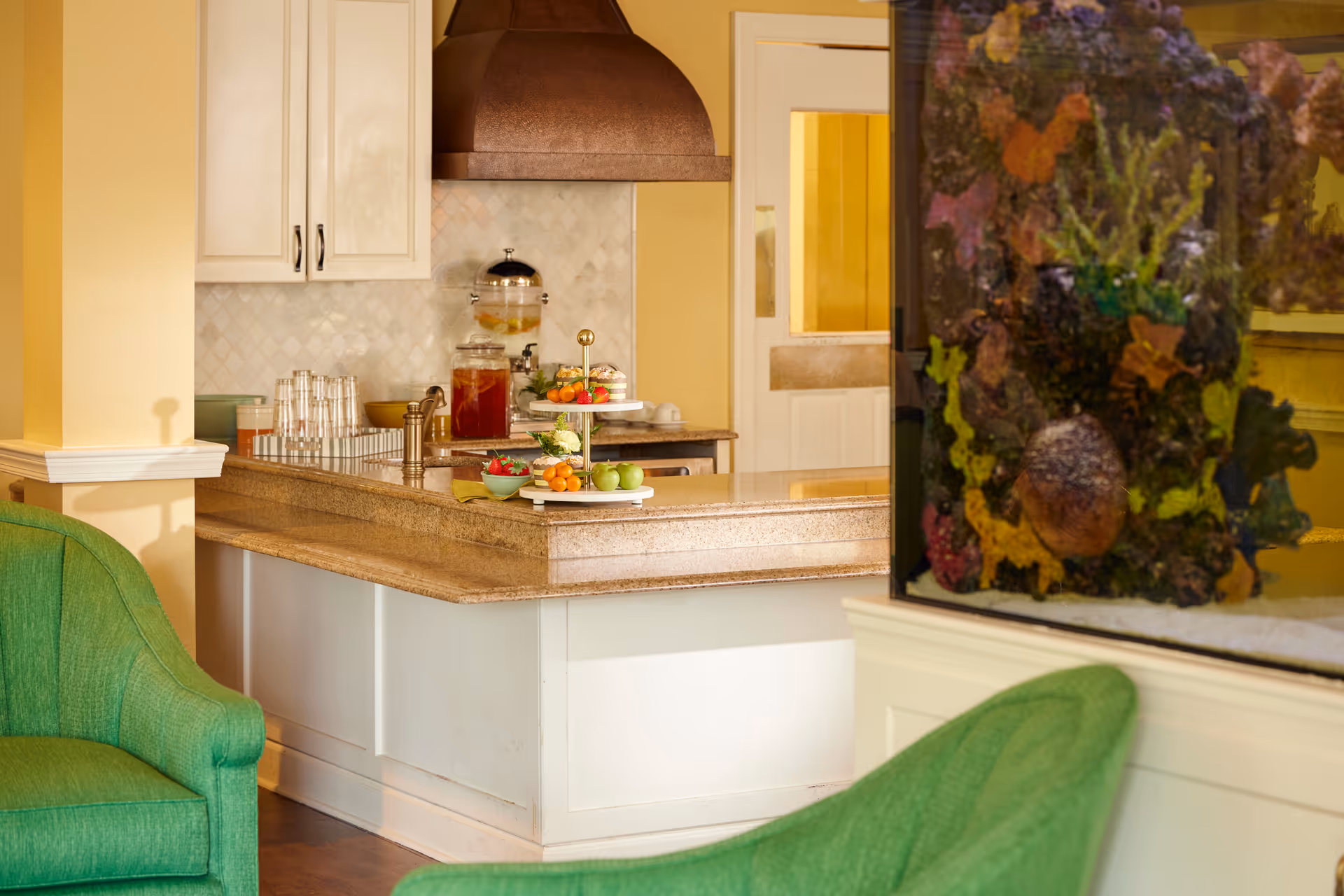 A cozy kitchen area with a granite countertop island, featuring a tiered tray with fruits and snacks, a beverage dispenser, and stacked glasses. The room has yellow walls, white cabinetry, a copper range hood, and green upholstered chairs. An aquarium with colorful coral and marine decorations is visible on the right side.
