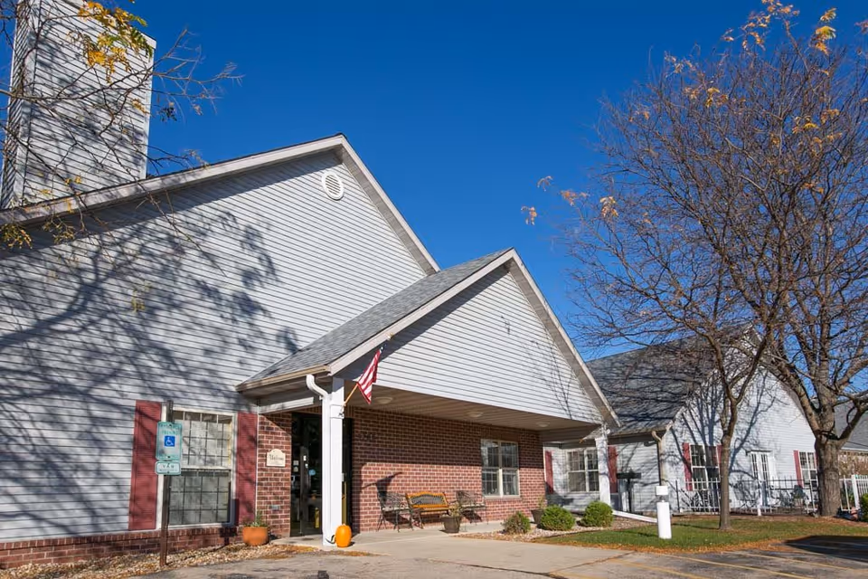 Exterior view of McFarland Villa Assisted Living building with a covered entrance, an American flag, a bench, and some landscaping including trees and shrubs under a clear blue sky.
