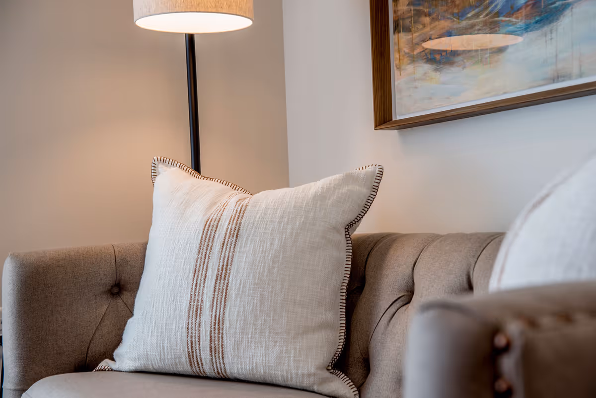 Close-up of a tufted sofa with a large decorative throw pillow, a floor lamp, and framed artwork on a neutral wall.