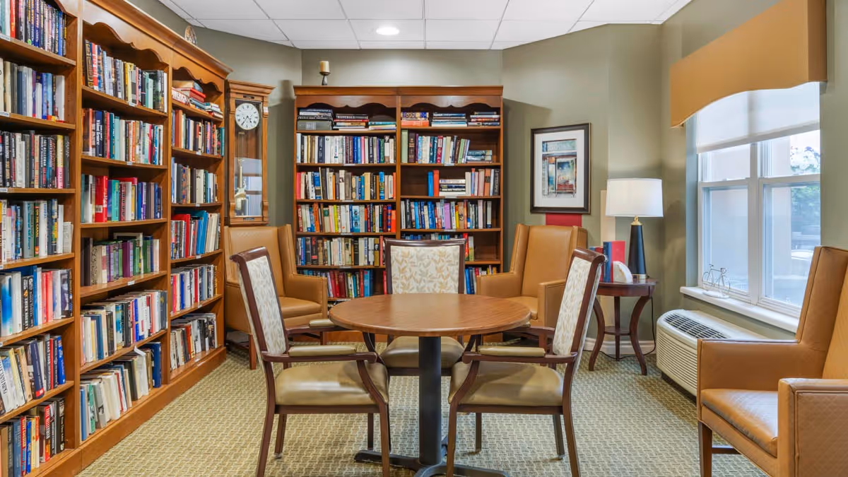 Cozy reading room with wooden bookshelves, a round table with chairs, armchairs, a lamp and a window.