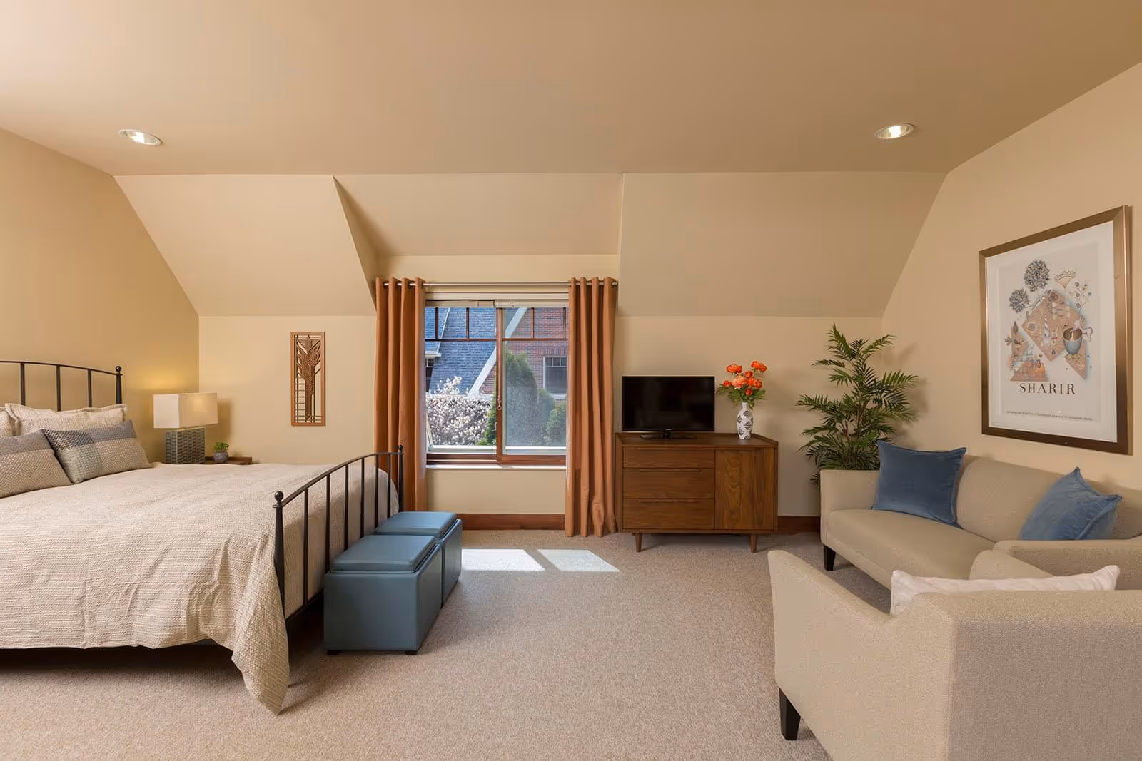 Well-lit bedroom with a metal bed, ottoman bench, window with curtains, wooden dresser with a TV, and a small seating area with sofa and armchair.