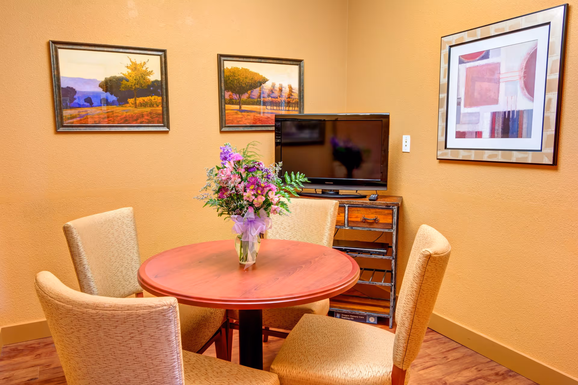 A small dining area with a round wooden table surrounded by four beige upholstered chairs. A vase with a colorful bouquet of flowers is placed in the center of the table. On the wall behind the table, there are three framed artworks and a flat-screen TV on a wooden stand.