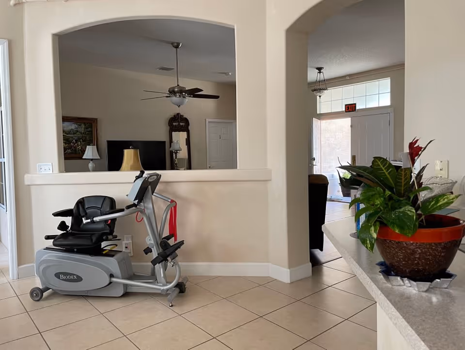 An indoor area of Brendlyn Assisted Living featuring a Biodex exercise machine on a tiled floor. The room has beige walls with an open archway leading to another room with a ceiling fan, a TV, a mirror, and a lamp. To the right, there is a countertop with a potted plant and an open door with an exit sign above it.