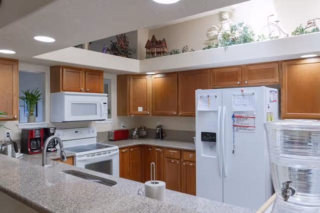 A clean and well-lit kitchen with wooden cabinets, a white refrigerator with notes attached, a white microwave above a white stove, a coffee maker, a toaster, and a water dispenser on the counter. There are decorative items on a ledge above the cabinets.