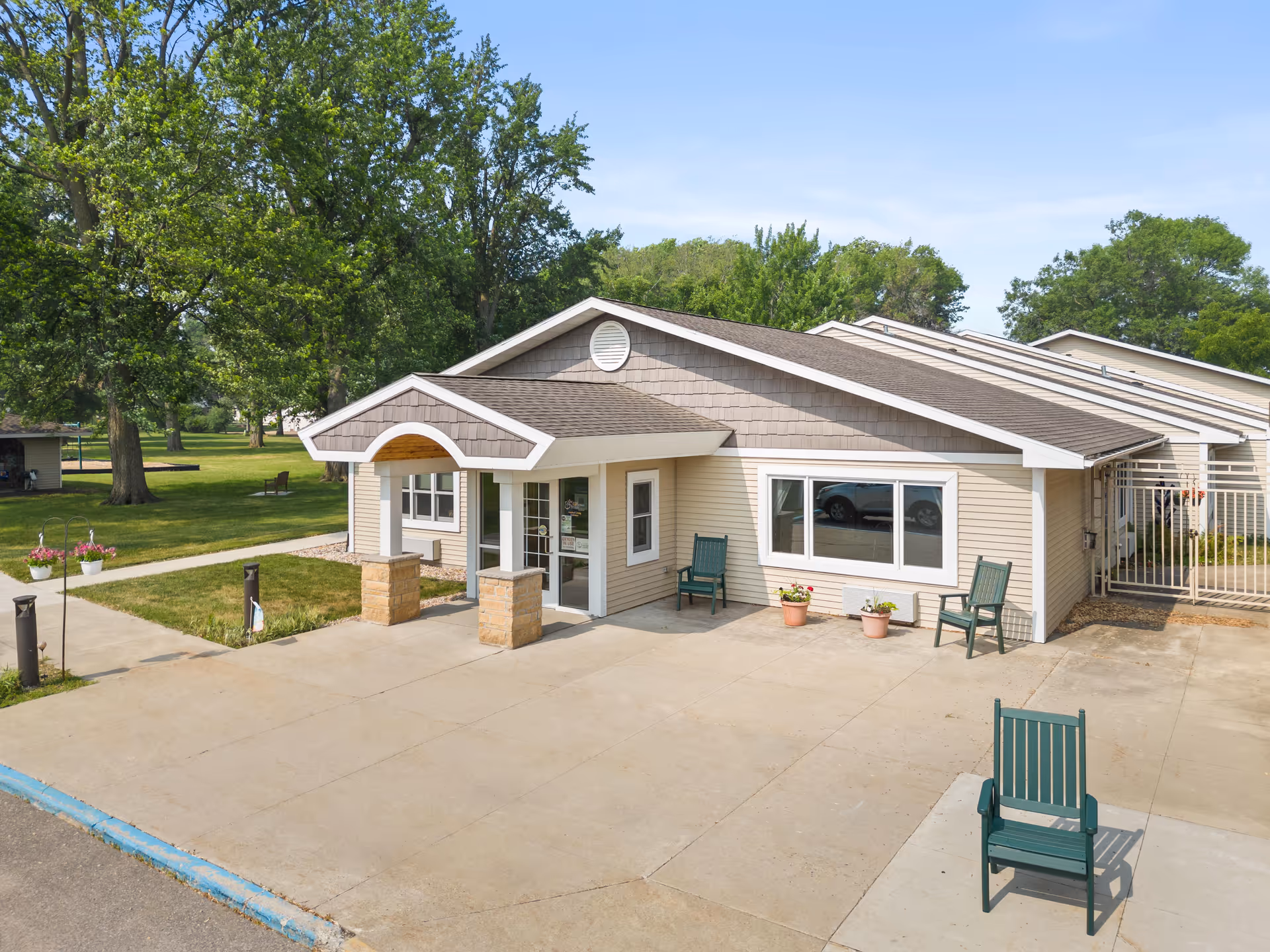 Front entrance of a single-story senior living facility with a covered porch, chairs, potted plants, and trees in the background.