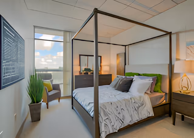 Sunlit bedroom with a four-poster bed, nightstands and lamps, a dresser, an armchair by a large window, and a potted plant.
