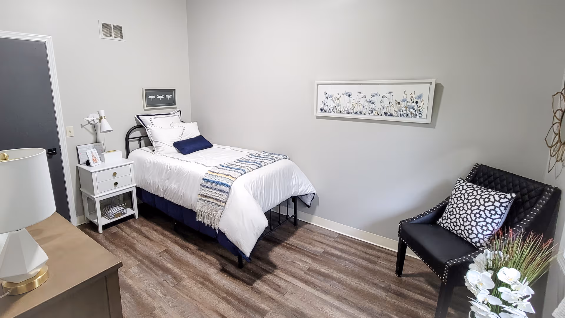 A neatly arranged bedroom with a single bed dressed in white bedding and a blue throw blanket. Next to the bed is a white nightstand with a lamp, picture frame, and books. A black chair with a patterned cushion is placed against the wall, which is decorated with a framed floral artwork. The floor is wood, and the walls are painted light gray.