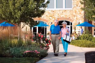 An elderly couple walking hand in hand on a paved pathway surrounded by landscaped gardens with flowers and trees. Behind them is a stone building with large windows and blue umbrellas shading outdoor seating areas.