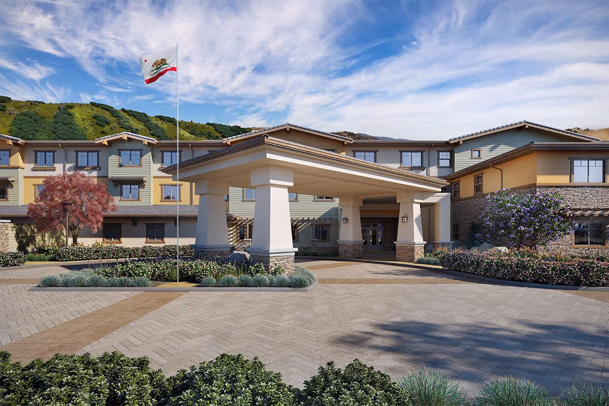 Front exterior view of a senior living facility named Sage Mountain with a covered entrance, landscaped greenery, a flagpole flying the California state flag, and a backdrop of green hills under a partly cloudy sky.
