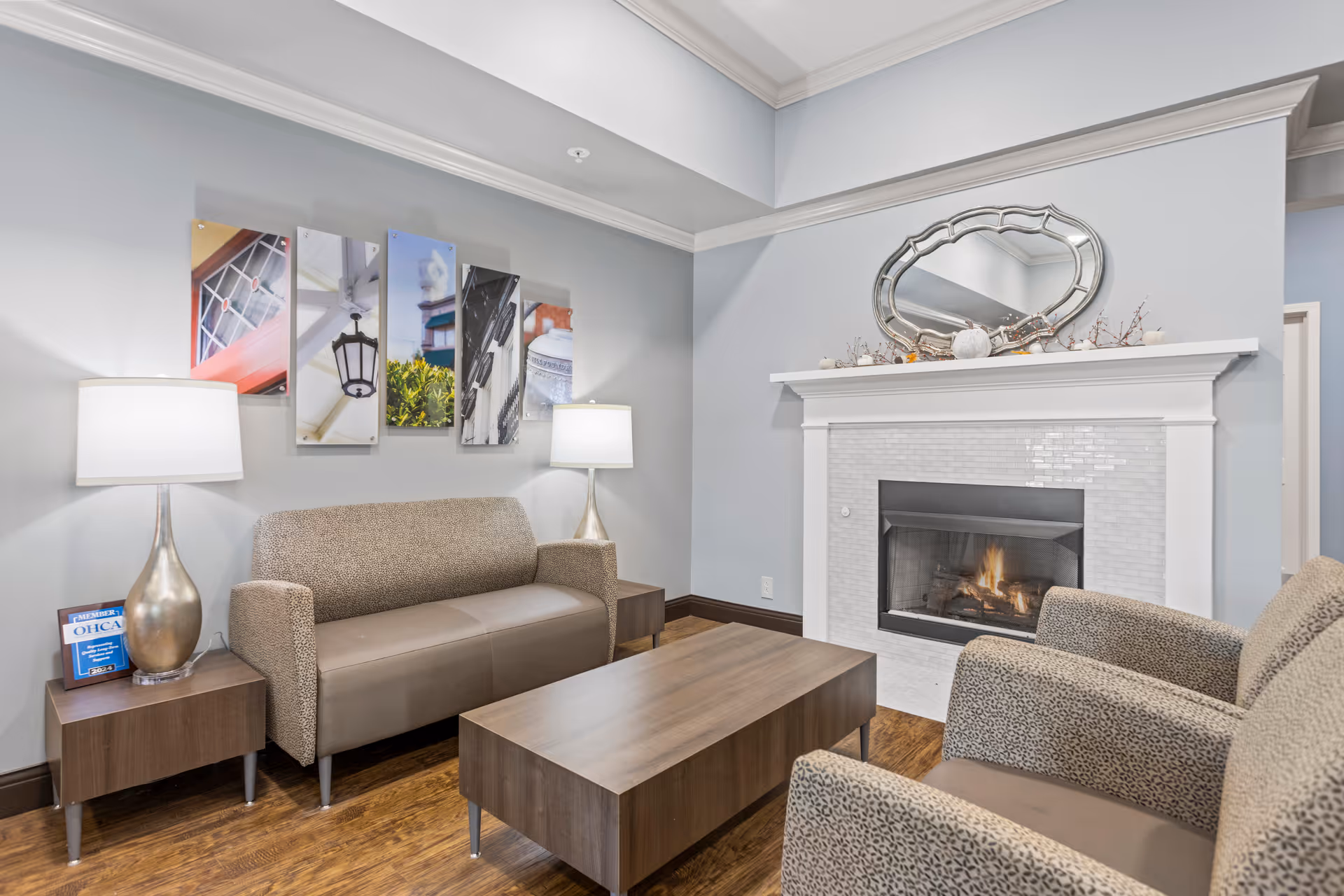 A cozy living room area with a beige patterned sofa and two matching armchairs around a wooden coffee table. The room features a white fireplace with a fire burning inside and a decorative mirror above the mantel. Two table lamps with white shades sit on wooden side tables flanking the sofa. The walls are painted light gray and decorated with a multi-panel artwork above the sofa. The floor is wooden.