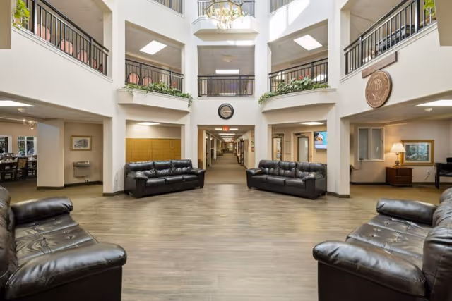 Spacious senior living community common area with four black leather sofas arranged around an open floor space. The area features a high ceiling with a chandelier, upper-level balconies with railings, and plants along the ledges. A hallway extends from the center, flanked by rooms and seating areas, with warm lighting and neutral-colored walls and flooring.