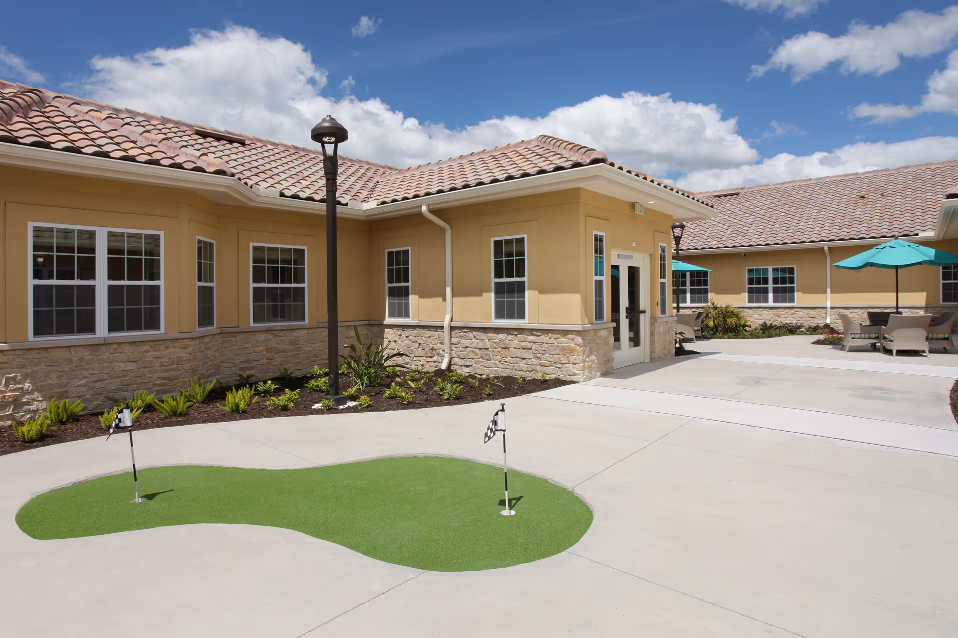 Outdoor courtyard area of The Fairways at Naples facility featuring a small putting green with two holes and checkered flags, surrounded by a concrete walkway. The building has beige walls with stone accents and a tiled roof. There are patio tables with turquoise umbrellas and chairs in the background under a partly cloudy blue sky.