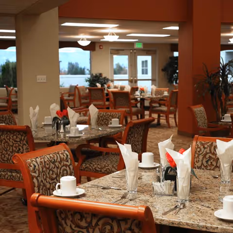 Dining area in a senior living facility with multiple tables set with white napkins, cups, and silverware. The room features patterned upholstered chairs, granite tabletops, warm lighting, and large windows with a view outside.