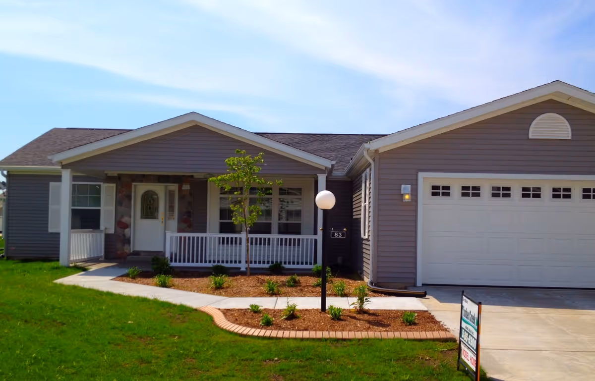 Front exterior view of a single-story house with gray siding, a white front door, a small porch with white railing, a young tree in the front yard, a driveway leading to a two-car garage, and a lawn with some landscaping.