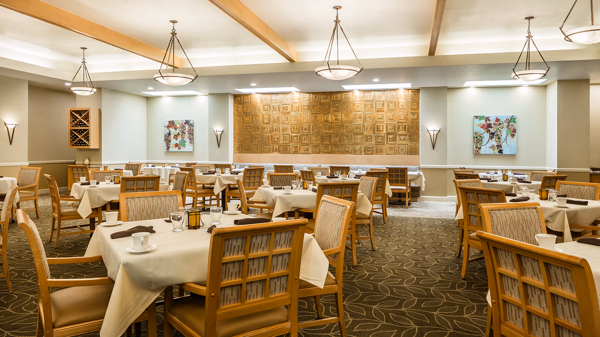 A spacious dining room with multiple tables covered in white tablecloths, each set with cups, glasses, and napkins. The room features wooden chairs with cushioned seats, decorative wall art, and warm lighting from ceiling fixtures and wall sconces. The carpet has a leaf pattern, and there is a wine rack on one wall.