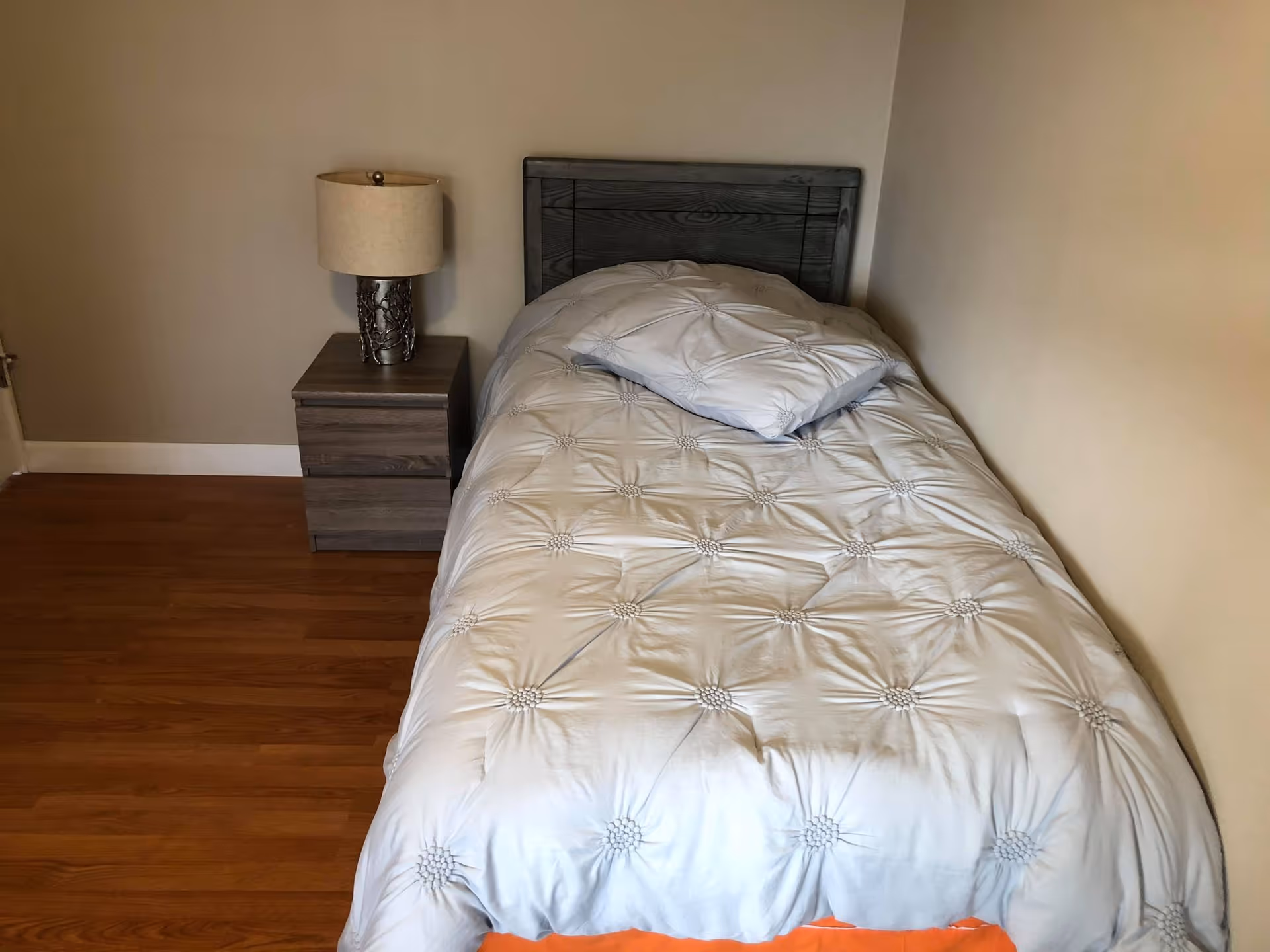 A small bedroom with a single bed against the wall. The bed has a white comforter with a floral tufted pattern and a matching pillow. Next to the bed is a wooden nightstand with two drawers and a decorative lamp with a beige lampshade. The floor is wooden and the walls are painted beige.