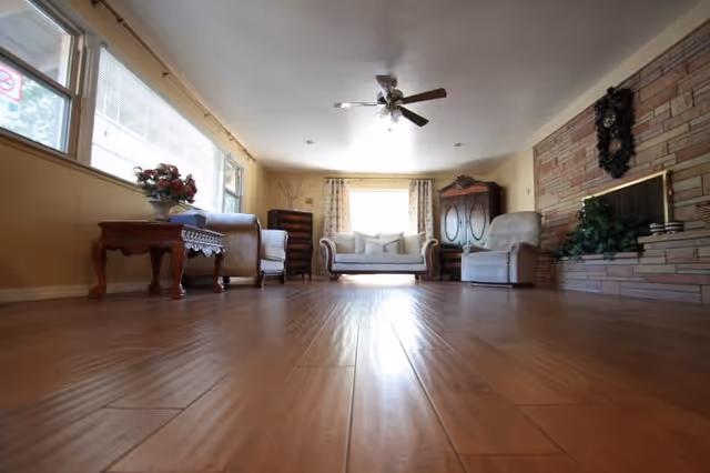 A spacious living room with wooden flooring, a ceiling fan, and large windows letting in natural light. The room is furnished with a sofa, armchairs, a wooden coffee table with a flower arrangement, a wooden cabinet, and a stone fireplace decorated with greenery and a wreath.