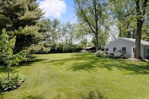 A spacious, well-maintained grassy yard with several mature trees and a small white building or shed on the right side under a partly cloudy blue sky.