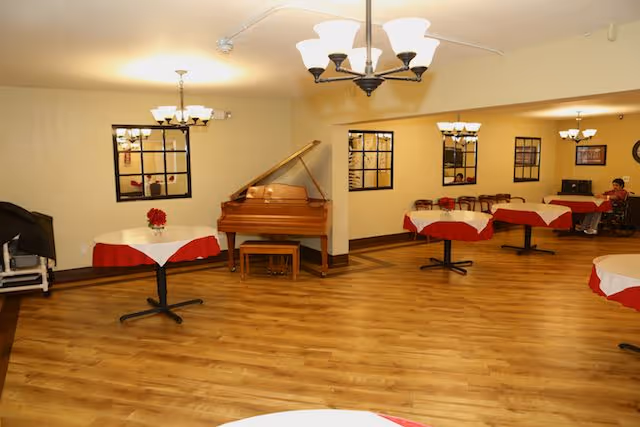 Interior view of a spacious room with wooden flooring, several round tables covered with white and red tablecloths, a grand piano against the wall, wall-mounted mirrors, and ceiling light fixtures. A person is seated at the far end of the room.