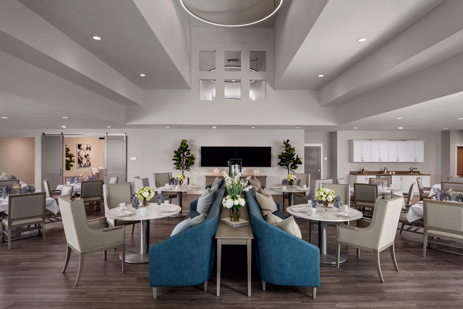 A spacious and modern dining room in a senior living facility with multiple round tables set with white tablecloths, floral centerpieces, and napkins. The room features comfortable chairs, blue cushioned bench seating in the center, wood flooring, and a high ceiling with recessed lighting. There are plants and a large flat-screen TV mounted on the far wall, with a kitchenette area visible in the background.