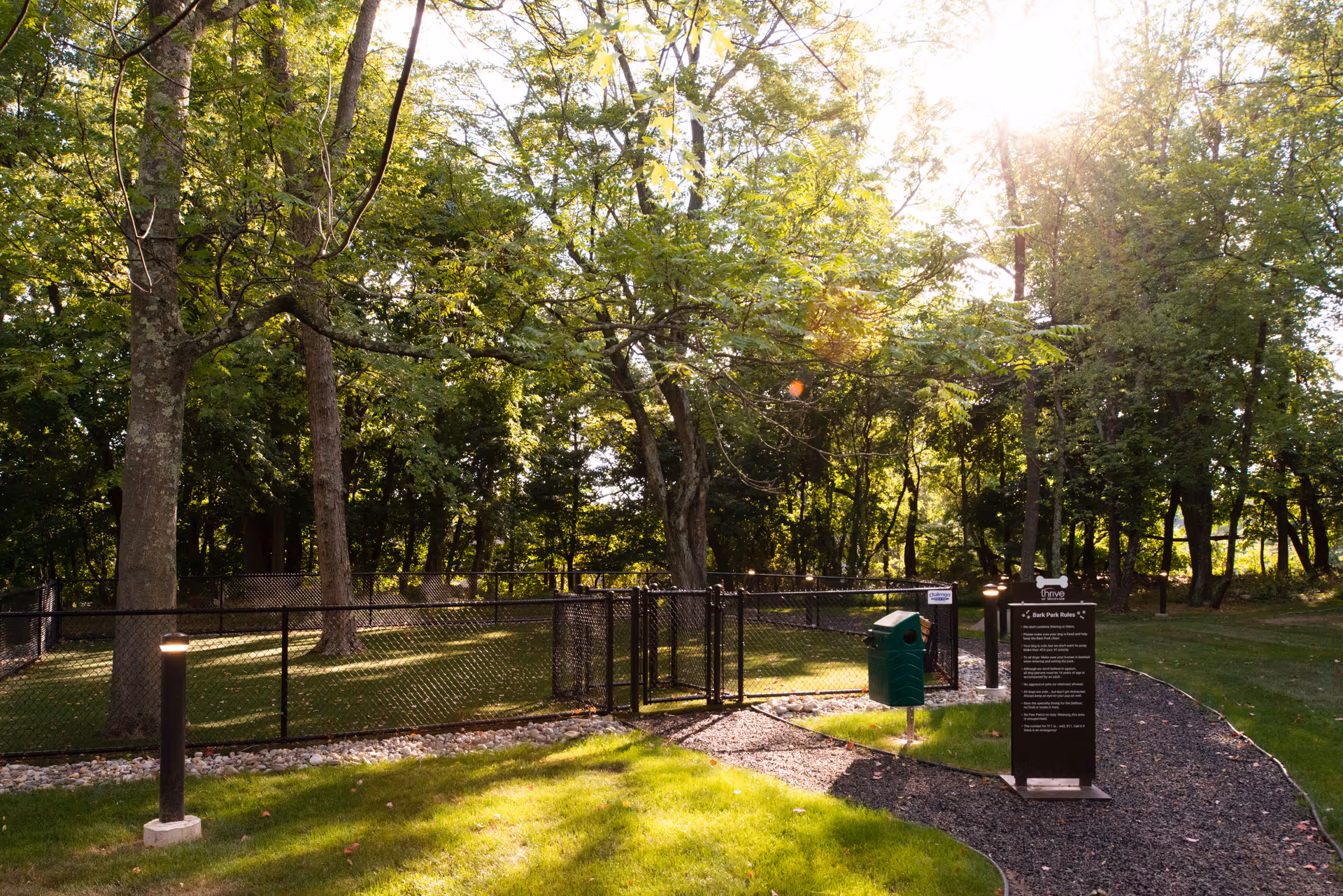 A fenced outdoor area in a senior living facility with trees and sunlight filtering through the leaves. There is a green trash bin and a sign with rules near the entrance gate, surrounded by a gravel pathway and well-maintained grass.