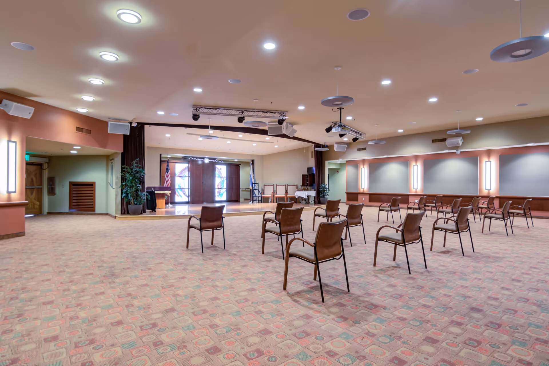 Large multipurpose activity room with spaced chairs facing a small stage and ceiling-mounted projectors.