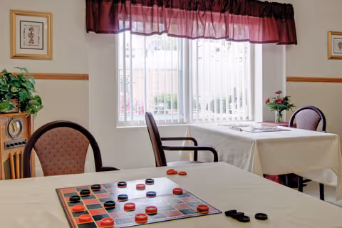 A bright room with two tables covered with white tablecloths. One table has a checkers game set up with red and black pieces. The room has three chairs with patterned upholstery, a window with vertical blinds and a maroon valance, a wooden cabinet with a plant on top, and framed artwork on the walls.