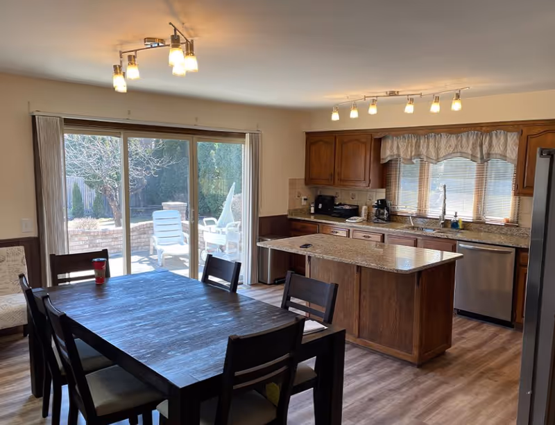A bright kitchen and dining area with wooden cabinets, granite countertops, and a kitchen island. The dining table has six chairs around it, and there is a sliding glass door leading to an outdoor patio with white chairs. The room has wood flooring and modern ceiling lights.