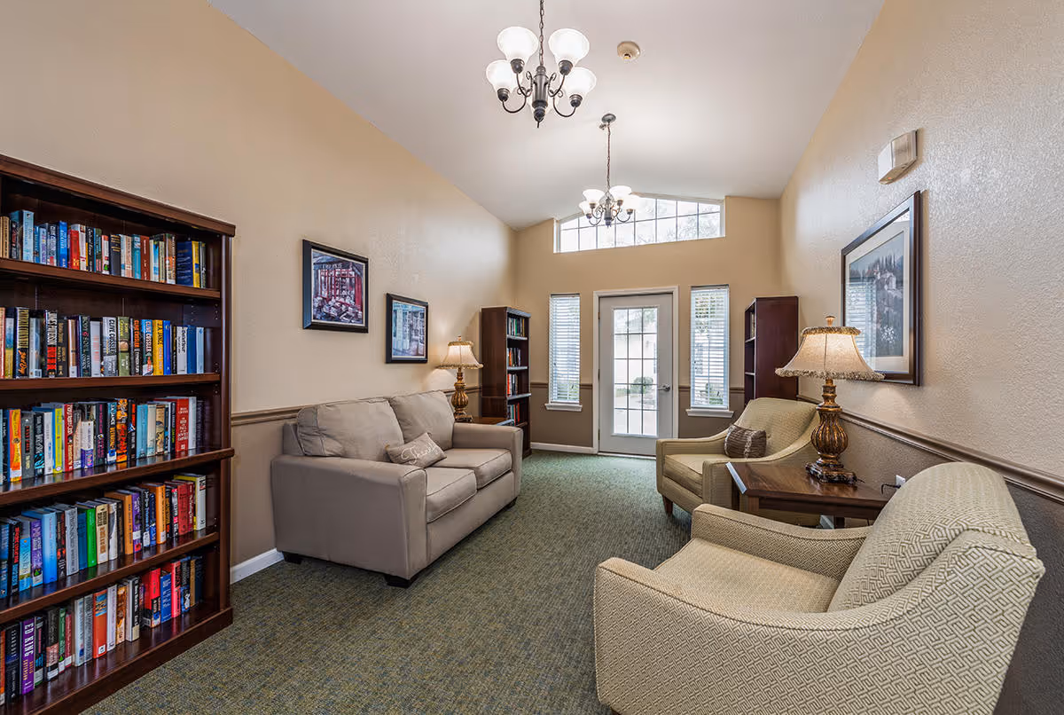 A cozy interior common area with beige walls and carpeted floor. The room features a beige sofa, two beige armchairs, wooden bookshelves filled with books, framed artwork on the walls, two table lamps on wooden side tables, and two chandeliers hanging from the ceiling. A glass door with windows on either side leads outside.