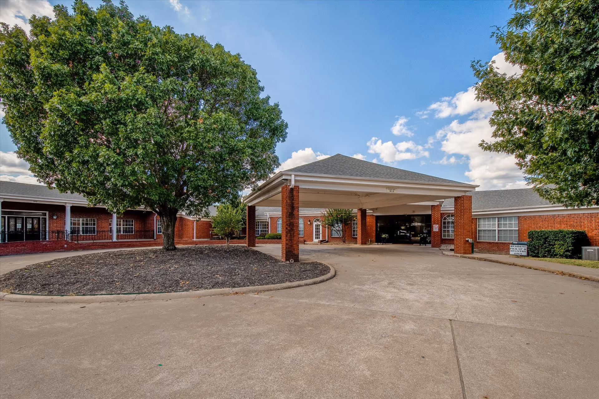 Front entrance of a red-brick building with a covered porte-cochere, circular driveway, and large trees.