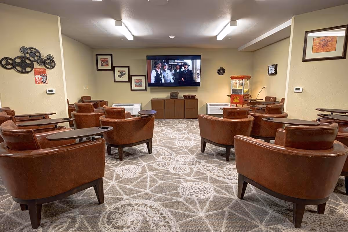 A cozy media room with multiple brown leather chairs arranged in rows facing a large flat-screen TV mounted on the wall. The room has a patterned carpet, framed pictures on the walls, and a popcorn machine in the corner.