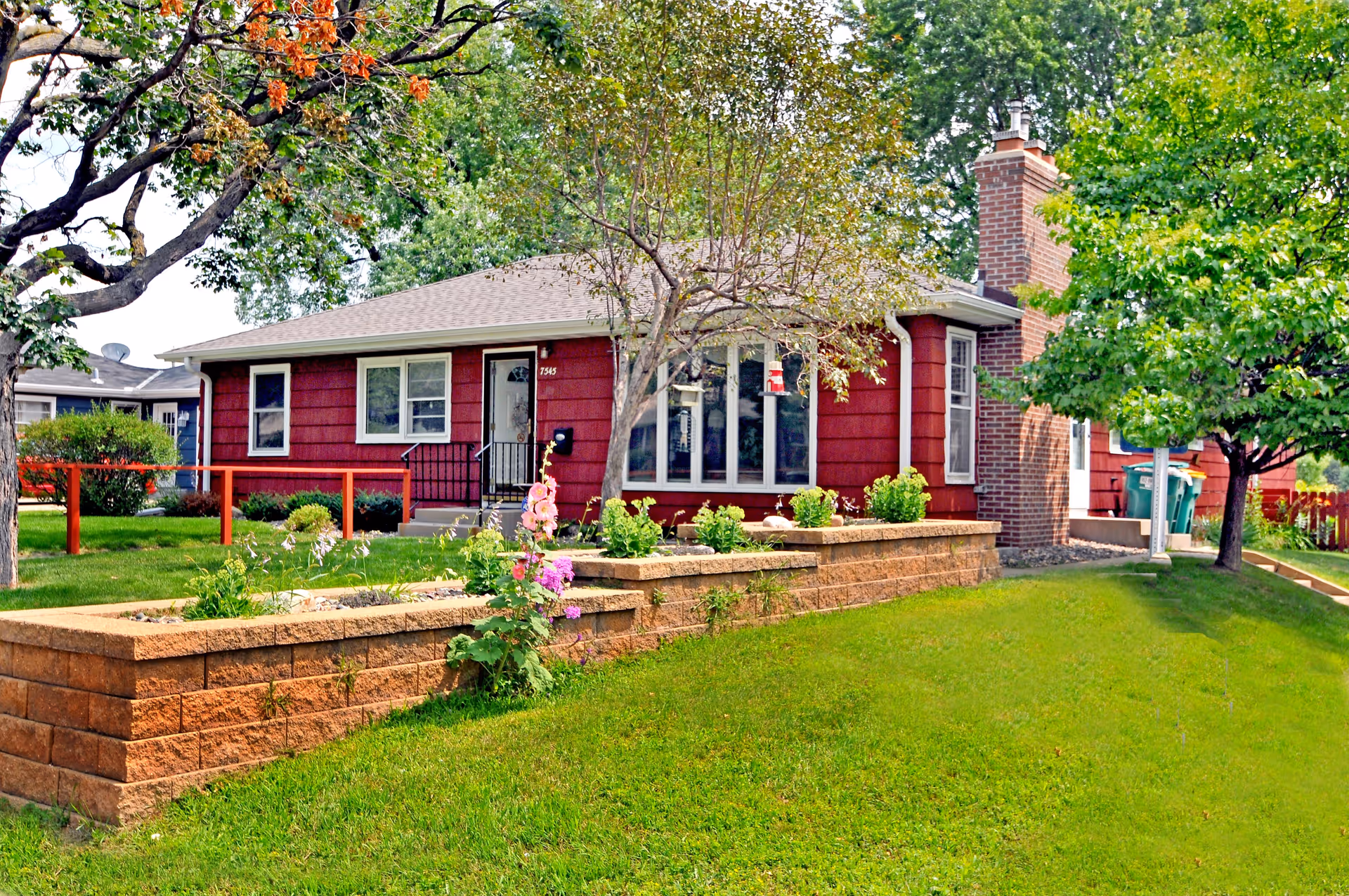 A single-story red house with white trim surrounded by green grass and trees. There is a brick chimney on the right side and a raised flower bed with various plants and flowers in front of the house. The house has several windows and a front door with steps leading up to it.