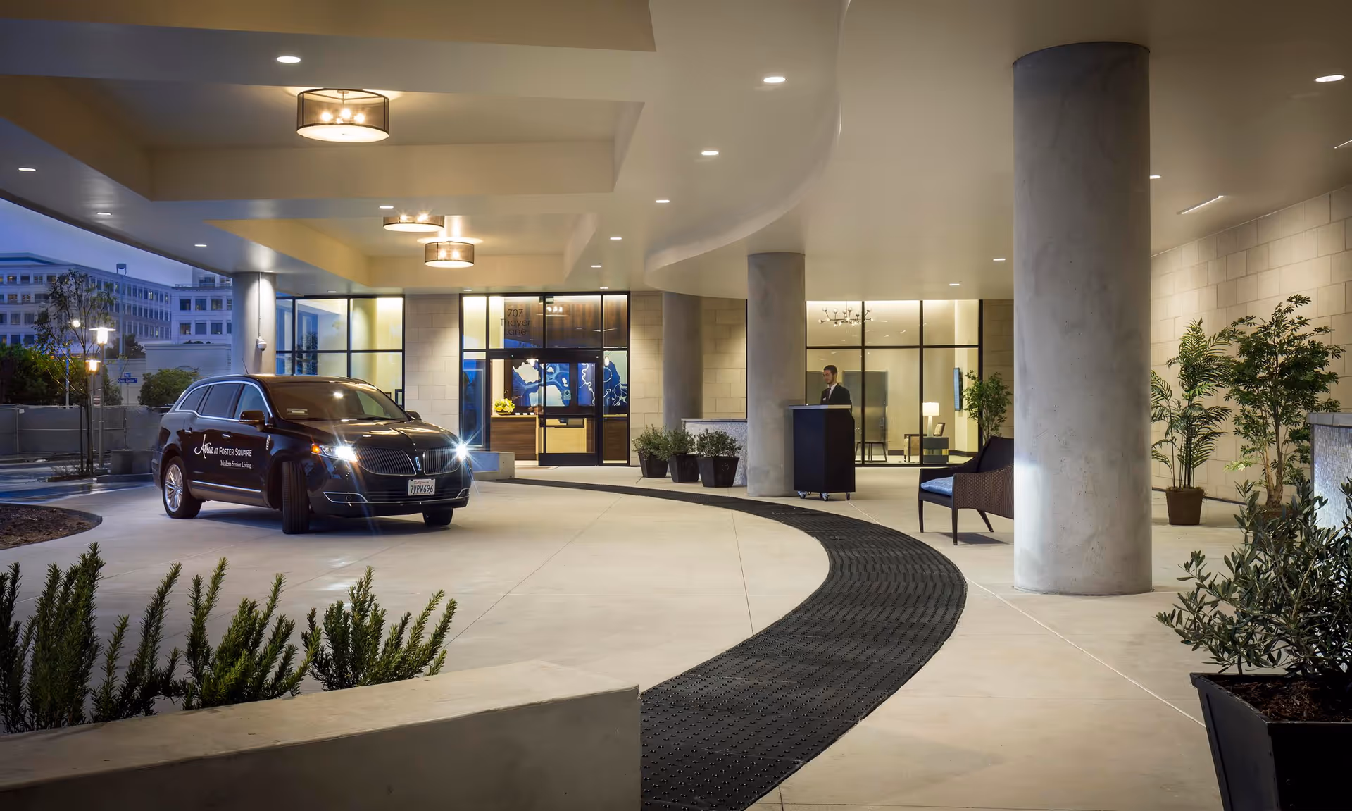 Covered entrance area of Atria At Foster Square with a black vehicle parked near the entrance. The area is well-lit with ceiling lights and features large concrete pillars, potted plants, and a person standing near a podium inside the building.