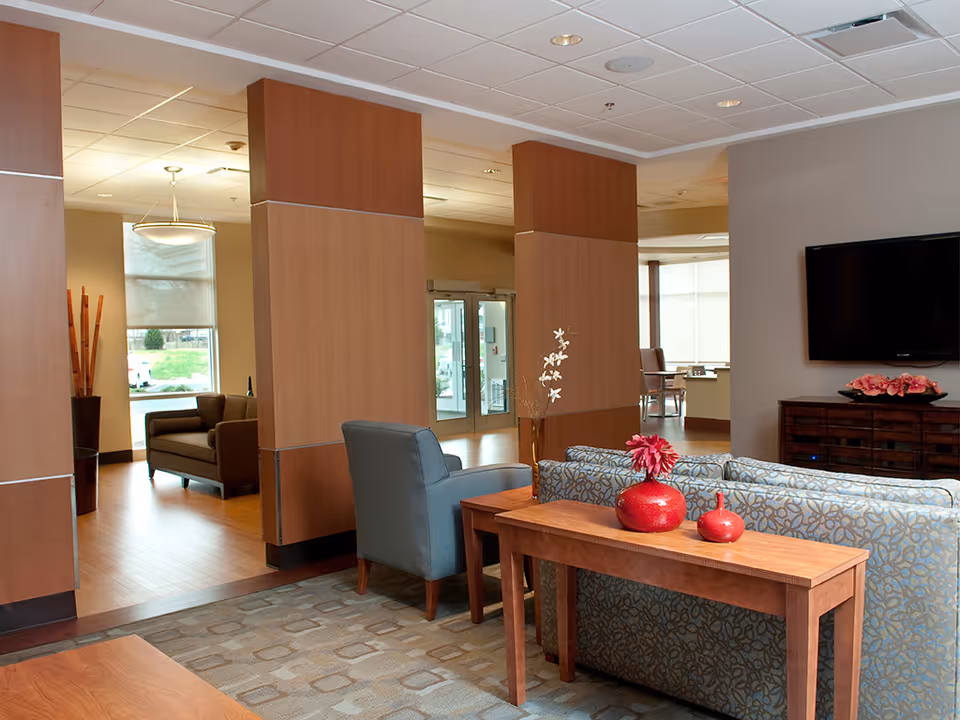 A modern and spacious living room area in BridgePort Healthcare Center featuring comfortable seating including a patterned sofa, a blue armchair, and a brown armchair. There is a wooden console table behind the sofa with red decorative vases and flowers. The room has wooden flooring, large windows allowing natural light, and a flat-screen TV mounted on the wall above a wooden cabinet with more pink flowers on top.