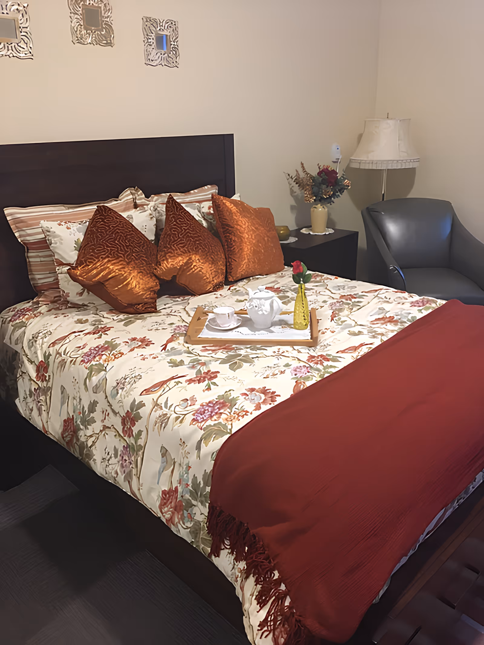 A decorated bedroom with a floral bedspread, copper-colored pillows, a red throw, a tea tray on the bed, a bedside lamp and an armchair.