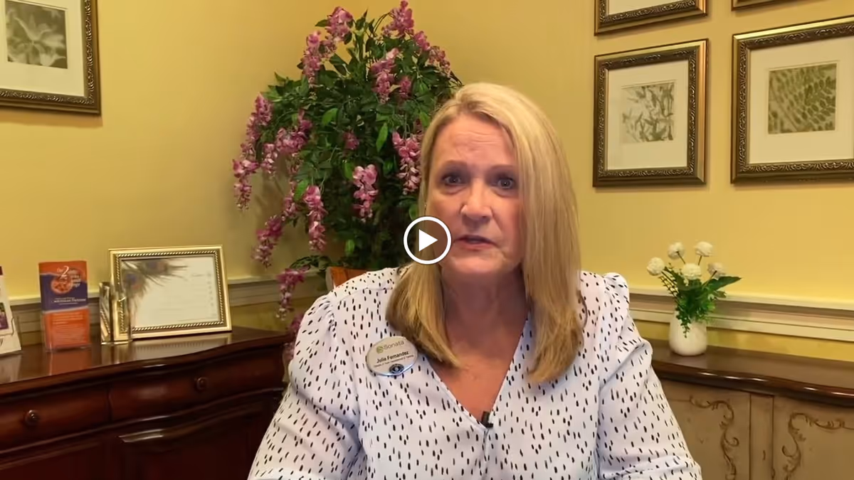 A woman with blonde hair wearing a white blouse with black dots and a name tag sits in a room with yellow walls. Behind her is a wooden sideboard with framed pictures, brochures, a vase with white flowers, and a large plant with purple flowers. The wall has three framed botanical prints.