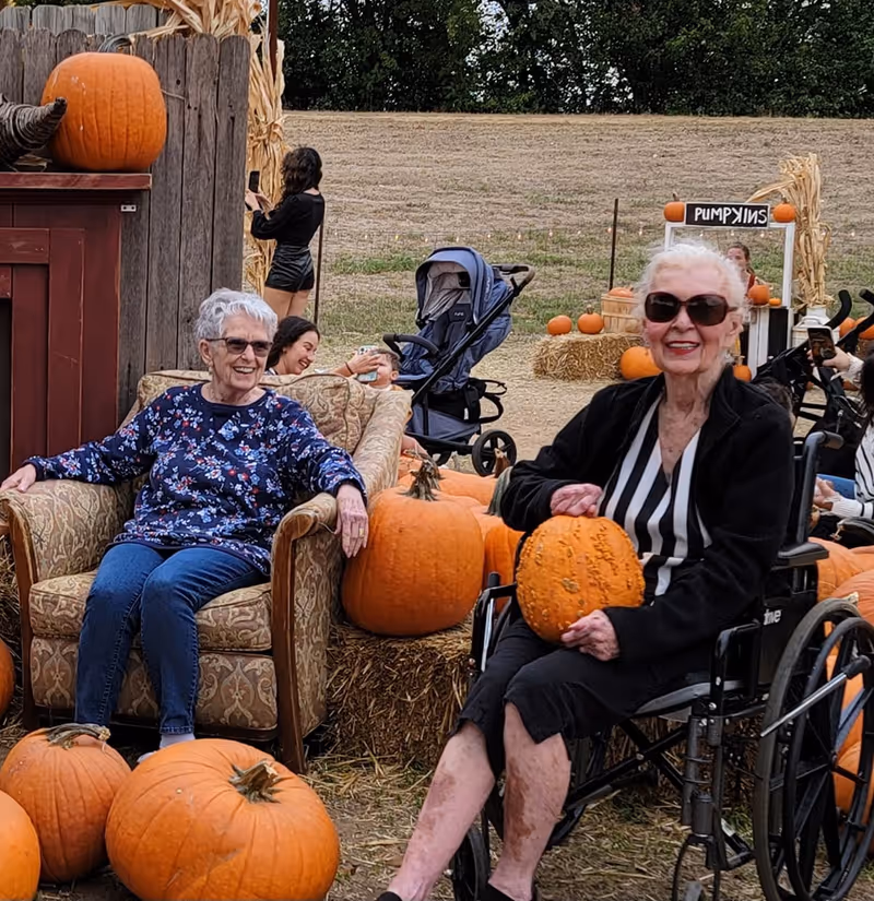 Two elderly women enjoying a pumpkin patch outdoors. One woman is sitting on a cushioned armchair, smiling and wearing a floral sweater and glasses. The other woman is in a wheelchair, holding a pumpkin and wearing sunglasses and a black and white striped top. Several pumpkins and hay bales surround them. In the background, a young woman is taking a photo with her phone, and a stroller is visible. A sign reading 'PUMPKINS' is also seen among the pumpkins and hay bales.