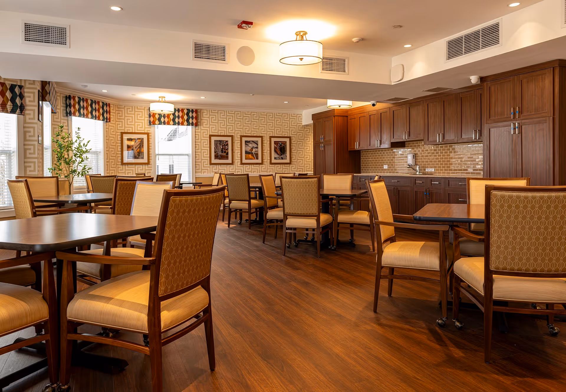A well-lit dining room with multiple wooden tables and cushioned chairs arranged neatly. The room features wooden flooring, patterned wallpaper, three framed pictures on the wall, and large windows with patterned valances. There are wooden cabinets and a countertop along one wall, with ceiling lights providing warm illumination.