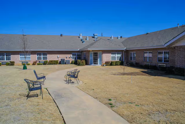 Single-story brick senior living building with a central entrance and a paved walkway through a grassy courtyard with metal benches.