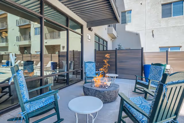 Outdoor patio area with four cushioned chairs arranged around a round fire pit table. There is a small white side table in the foreground. The patio is enclosed with privacy fencing and adjacent to a building with large windows reflecting the outdoor furniture.
