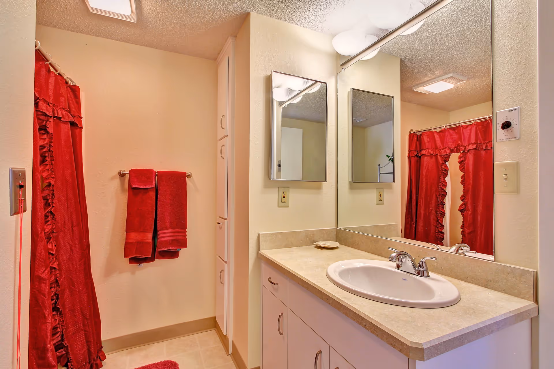 A bathroom with a beige countertop and white sink, a large mirror above the sink, two smaller mirrors on the adjacent wall, red towels hanging on a towel rack, and a red shower curtain with ruffles. The walls are light-colored and there is a ceiling light fixture.