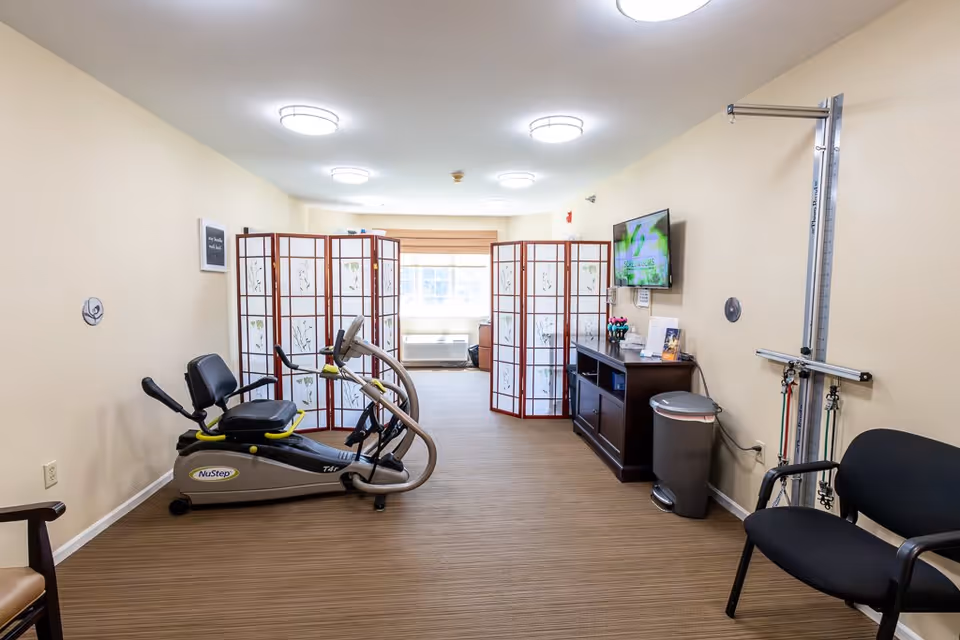 A small exercise room with a NuStep recumbent cross trainer, a black chair, a wooden cabinet with a mounted TV above it, and a folding room divider with floral patterns near a window.